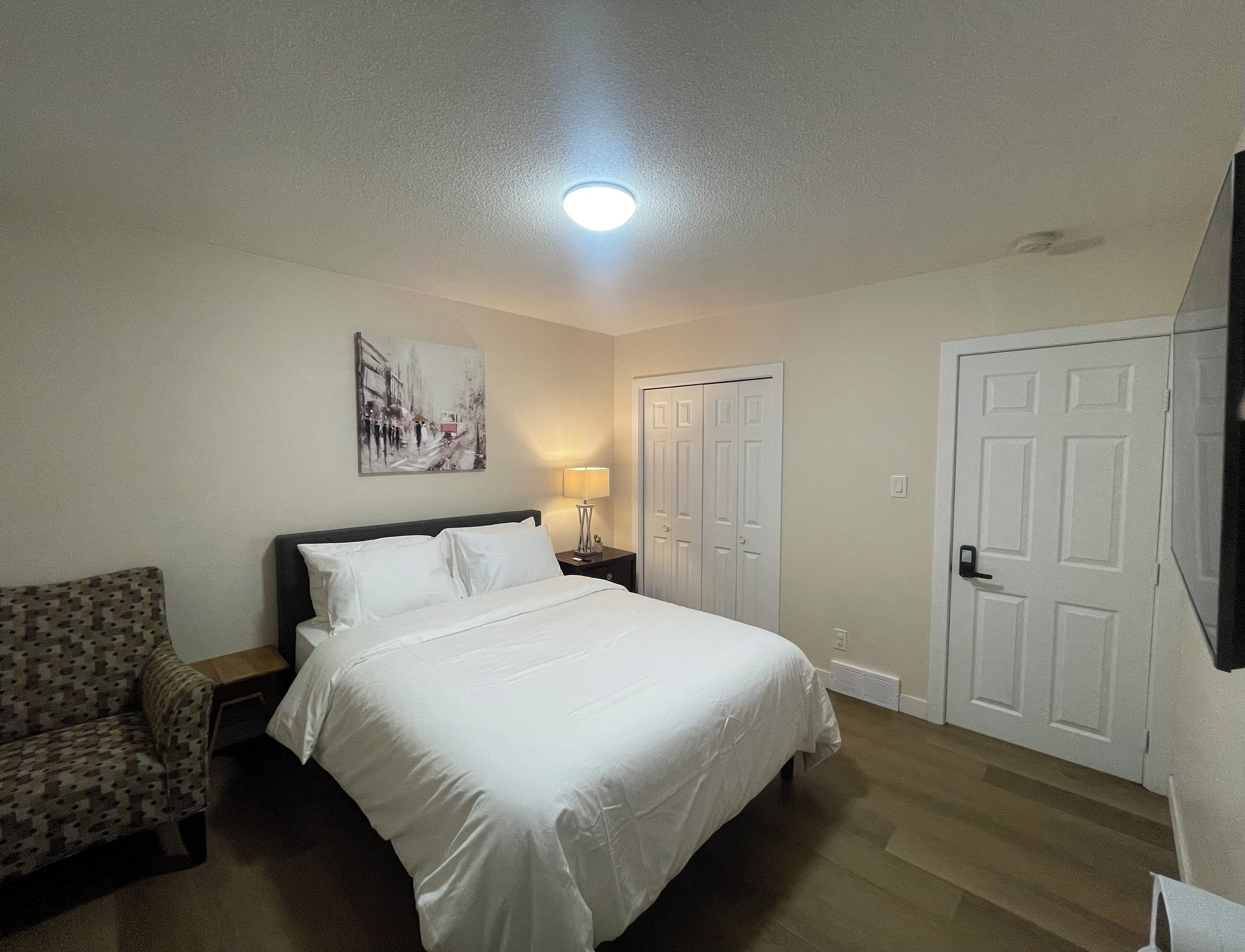 A hotel room with a bed, a chair, a nightstand, a lamp, a painting, and a door.