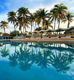 A pool area with palm trees, lounge chairs, and a covered seating area. - 2bd Beachfront Master Ste, Bay View w/balcony, Pool, nearby dining, beach access (Miami Beach)