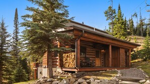 Une cabane en rondins dotée d’un porche, entourée d’arbres et d’un mur de pierres.