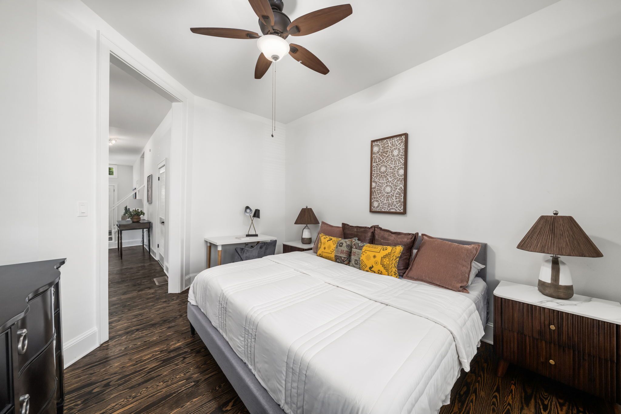 A bedroom with a large bed, a ceiling fan, bedside lamps, a nightstand, and a small table with a lamp.