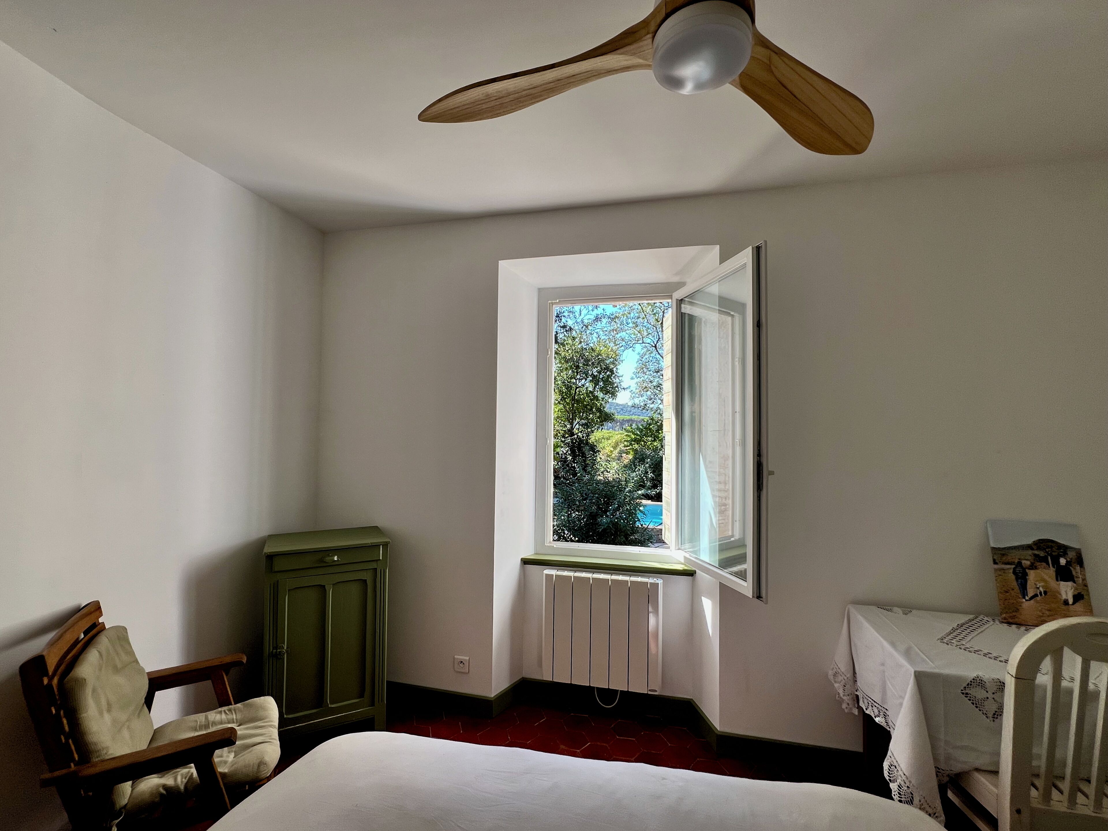 A room with a bed, a wooden chair, a table with a tablecloth, a radiator, and a window with a view of greenery and a building.