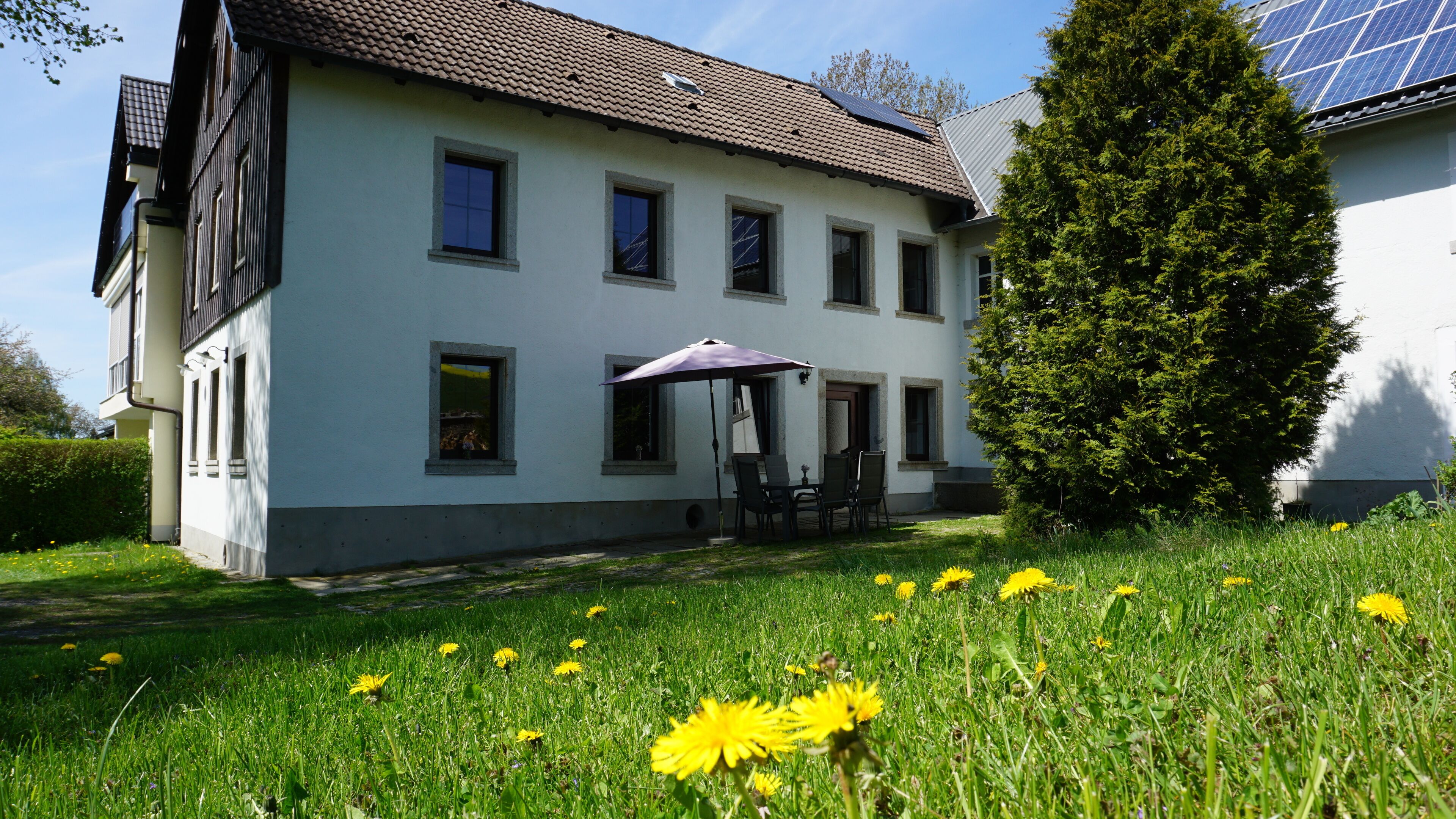 A two-story house with a patio area, a dining table with chairs, and a large umbrella.