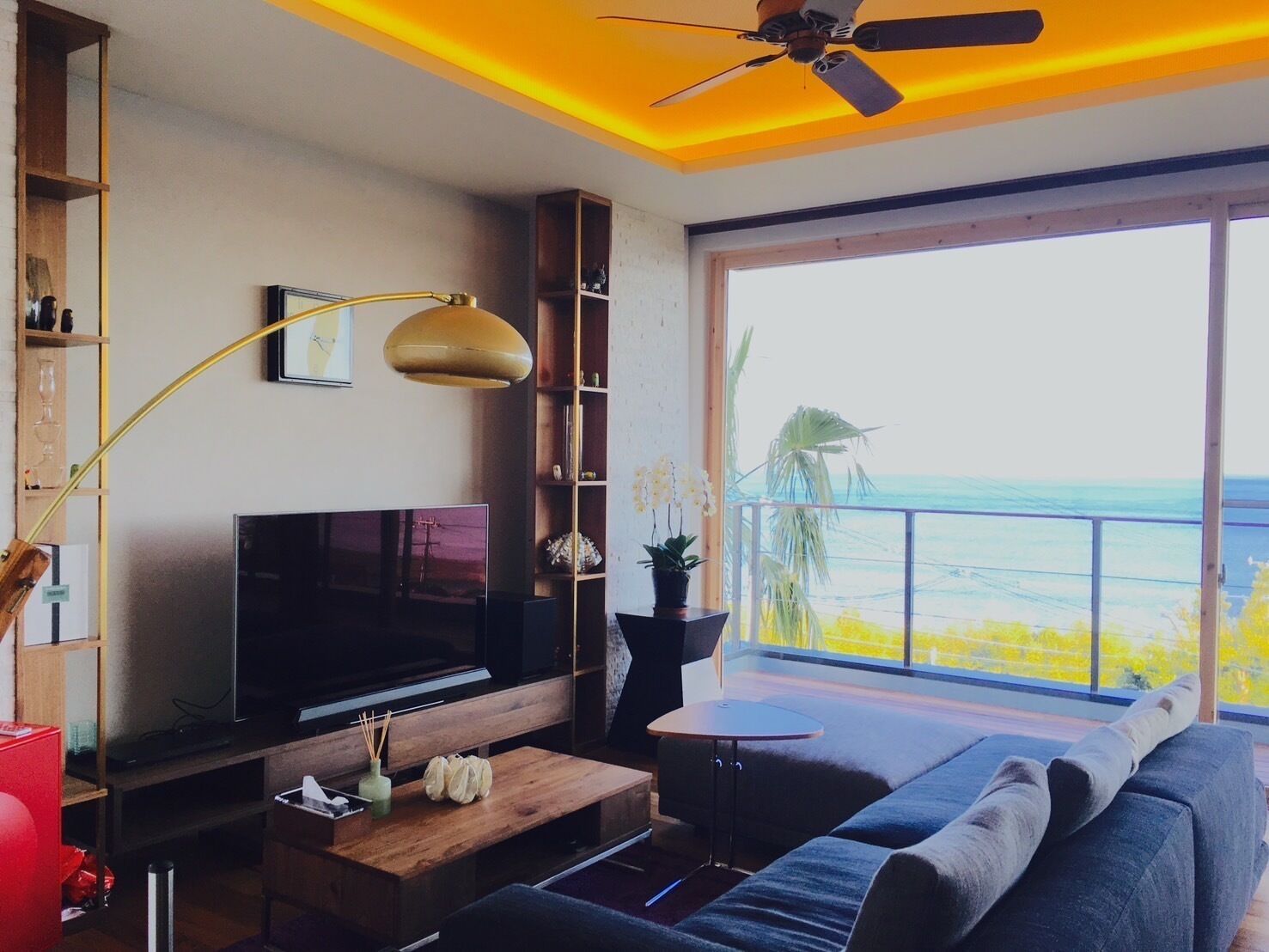 Un salon moderne avec un téléviseur, des meubles en bois, un ventilateur de plafond et une grande fenêtre donnant sur une plage.