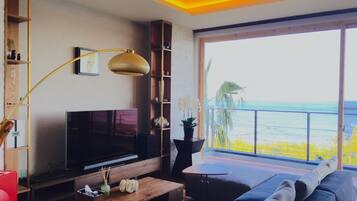 Un salon moderne avec un téléviseur, des meubles en bois, un ventilateur de plafond et une grande fenêtre donnant sur une plage.