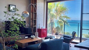 A modern living room with a large window overlooking the ocean, a ceiling fan, and a mix of furniture including a sofa and a coffee table.