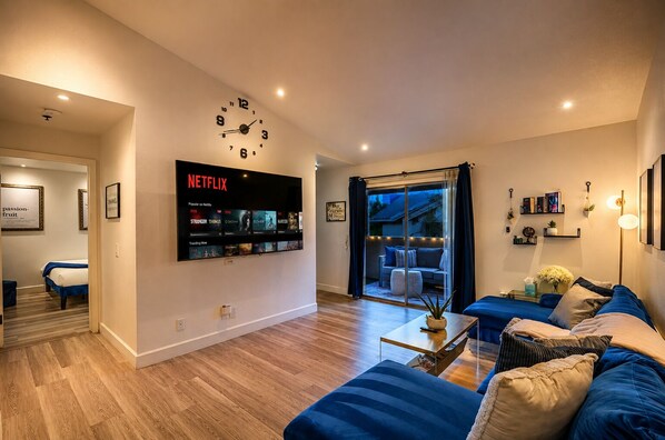 Un salón moderno con un televisor de pantalla plana mostrando Netflix, un reloj grande y una zona de estar acogedora con un sofá y una mesa de centro.
