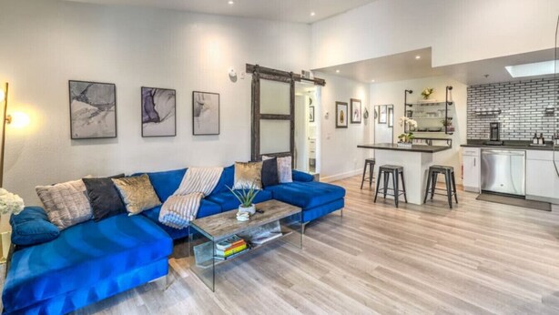Una sala de estar moderna con un sofá azul, una mesa de centro de vidrio y una zona de cocina con barra y armarios blancos.