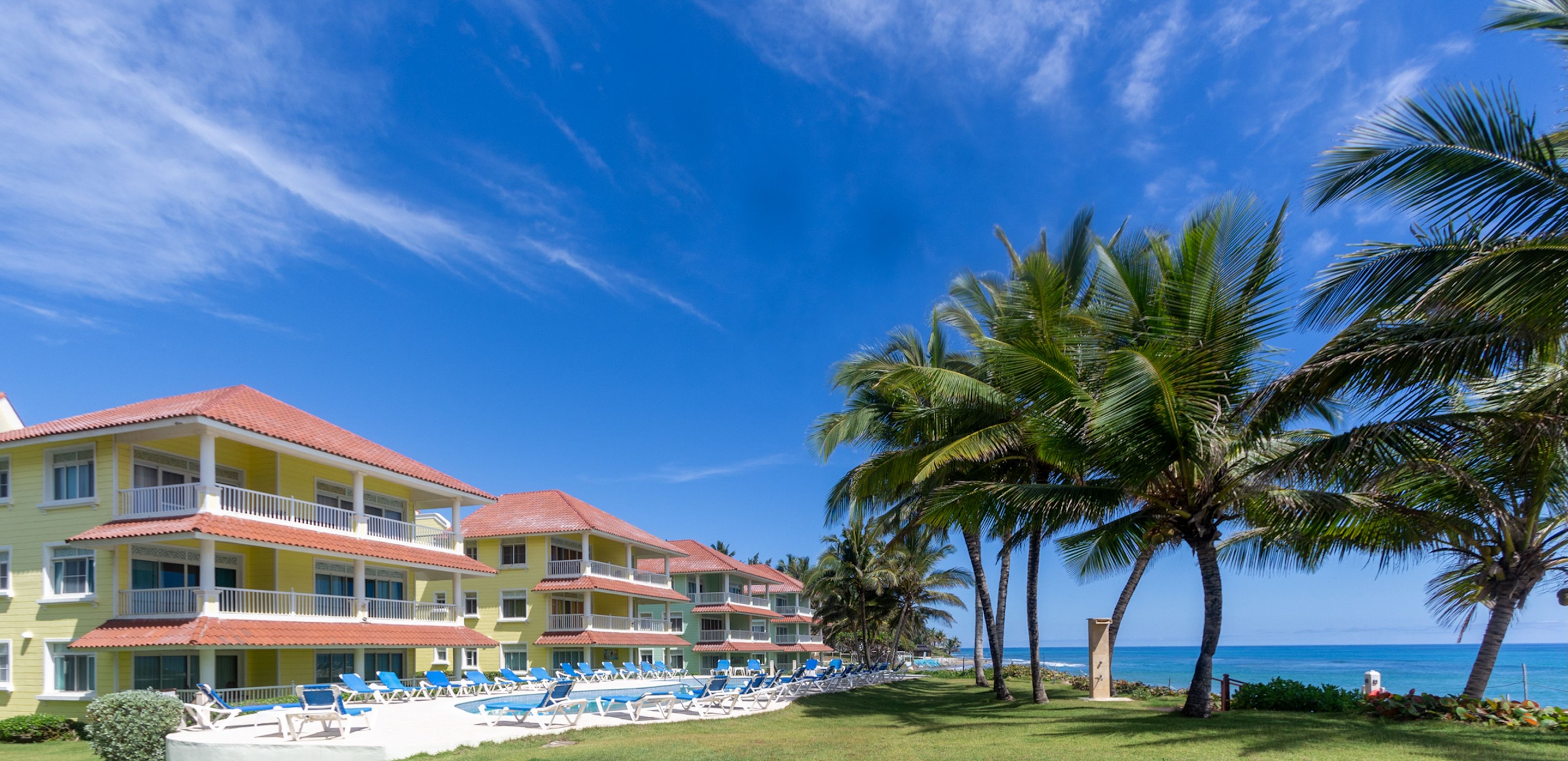 Un hotel frente a la playa con varios balcones, una zona de piscina con sillones de descanso y palmeras.