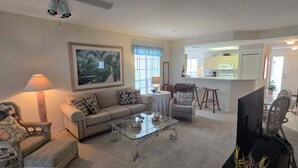 A living room with a sofa, armchairs, a coffee table, a TV, and a kitchen area in the background.