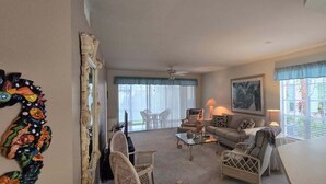 A living room with a sofa, armchairs, a coffee table, and a TV. There are windows with blinds, a ceiling fan, and a decorative wall piece.