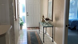 A hallway with a white door, a glass table with decorative items, a patterned rug, and a bed with a blue blanket in the adjacent room.