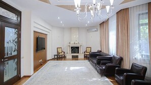 A spacious living room with a fireplace, leather sofas, a chandelier, and a large window with curtains. - Villa Kali Nadyrova (Almaty)