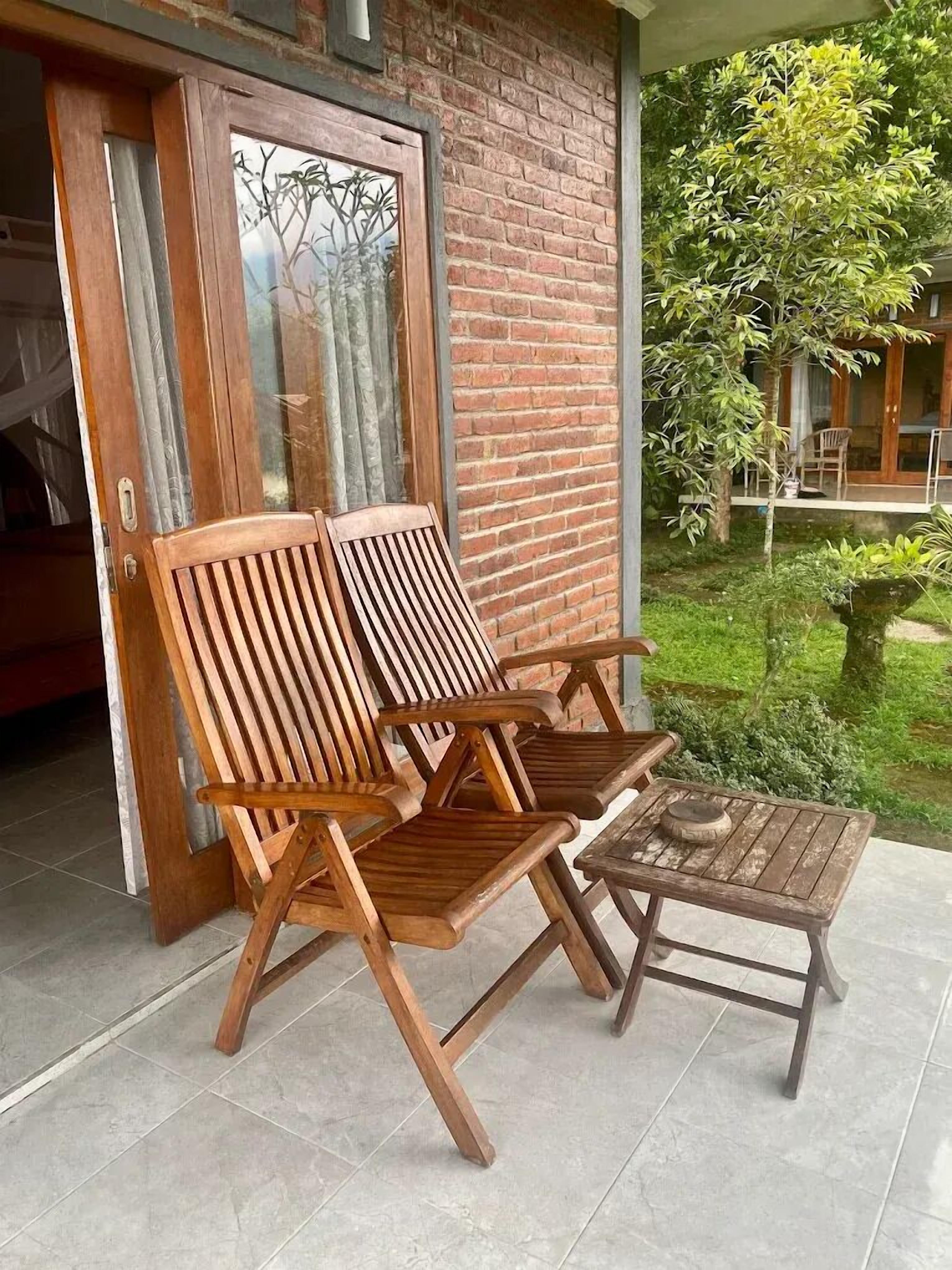 Una terraza de madera con dos sillones de descanso y una mesita, un muro de ladrillo y un ventanal de vidrio que da a un espacio cubierto.