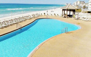A swimming pool with a curved edge, adjacent to a beach with lounge chairs and a gazebo.