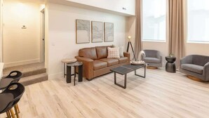 Un salon moderne avec un canapé en cuir marron, deux fauteuils, une table basse et une table d’appoint. Trois œuvres d’art encadrées ornent le mur, ainsi qu’une lampe sur pied et une grande fenêtre avec des rideaux.