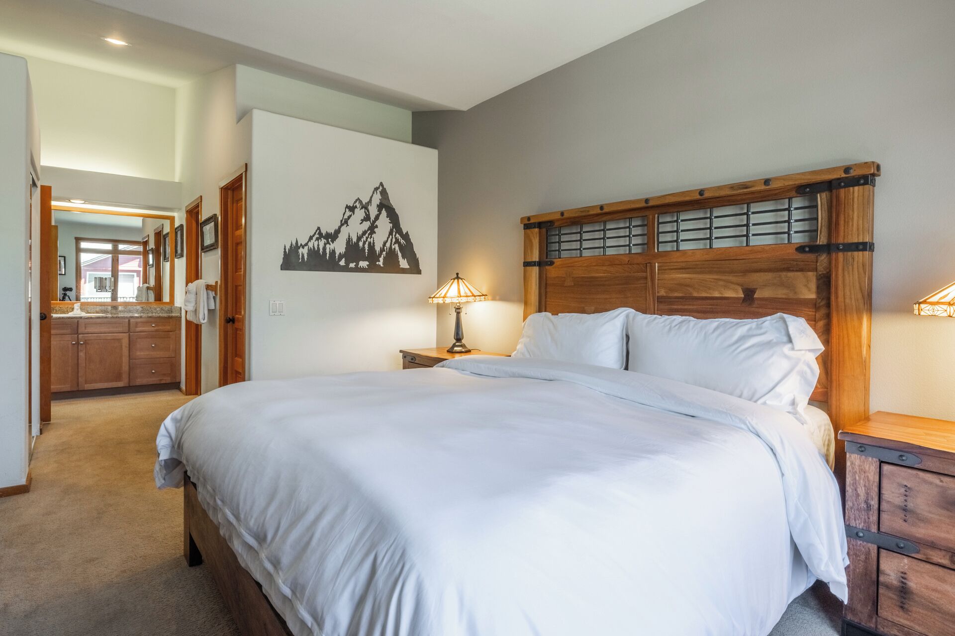 A bedroom with a large bed, wooden headboard, two bedside tables with lamps, a bathroom with a wooden vanity, and a wall-mounted picture of mountains.