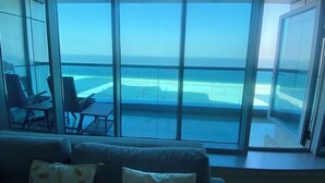 A room with a large glass wall offering a view of a beach and ocean. There is a sofa with cushions and a small table with chairs.