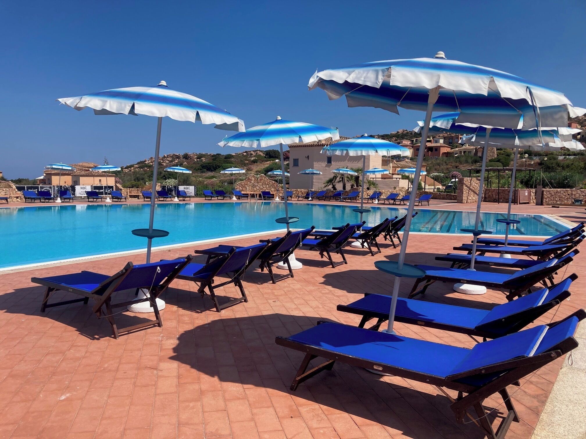 A poolside area with blue and white umbrellas, sun loungers, and a clear view of the pool.