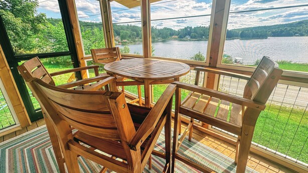 A wooden deck with a round table and chairs, overlooking a lake and forest.