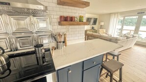 A modern kitchen with a black stove, blue cabinets, and a white countertop. There is a wooden shelf above the counter holding glasses and a TV. A living area with a sofa and a view of a lake is visible through large windows.