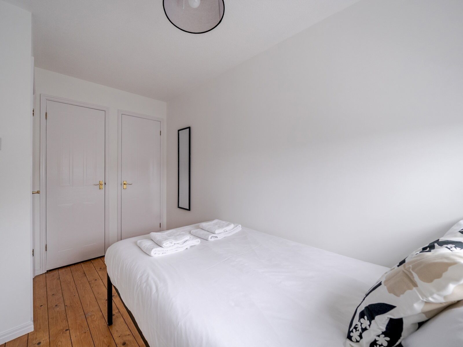 A bedroom with a large bed, a wooden floor, and white walls. There are two doors and a framed mirror on the wall.