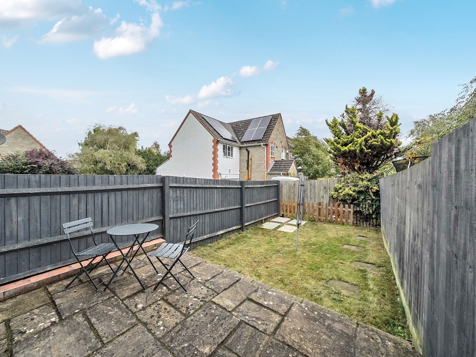 A backyard with a wooden fence, a small patio with two chairs and a table, and a house with solar panels in the background.