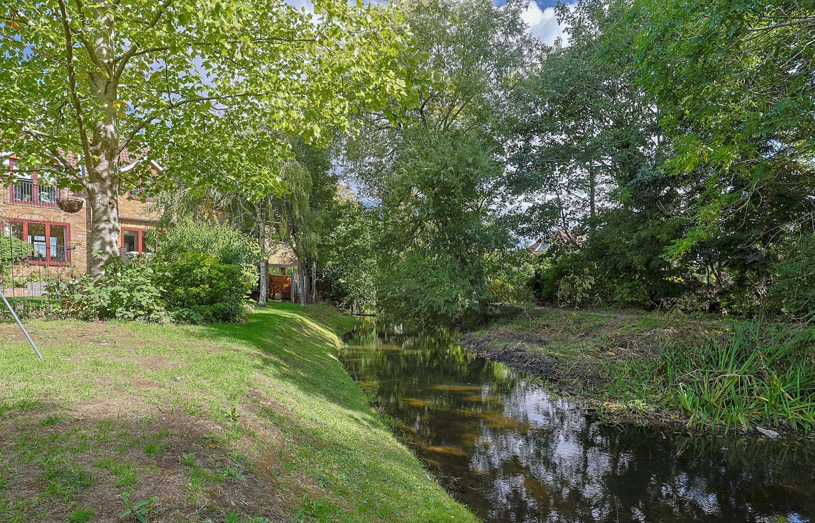 Een rustgevende rivieromgeving met een kleine brug, weelderige begroeiing en een gebouw met rode ramen op de achtergrond.