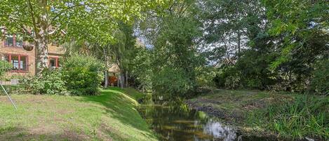 A serene riverside setting with a small bridge, lush greenery, and a building with red windows in the background.