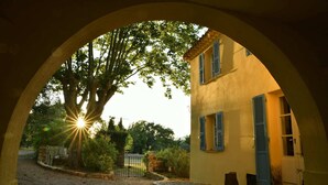 A rustic building with a yellow facade and blue shutters, viewed through an arched doorway with sunlight streaming through.
