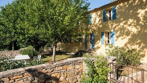 A stone wall, a yellow building with blue shutters, and a tree in a courtyard.