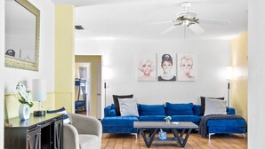 Una sala de estar con un sofá azul, una mesa de centro y un ventilador de techo. Hay lámparas de pared y cuadros enmarcados en la pared.