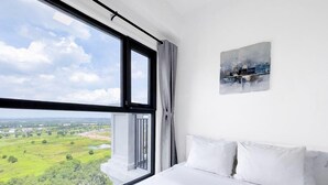 Un dormitorio con un ventanal grande, ropa de cama blanca y un cuadro en la pared.