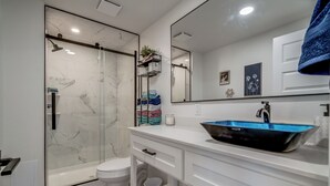 A modern bathroom with a glass shower enclosure, a white toilet, a white vanity with a black sink, and a large mirror. - Charming 1-bedroom cottage on historic Rt. 66 on the west side of Yukon. (El Reno)