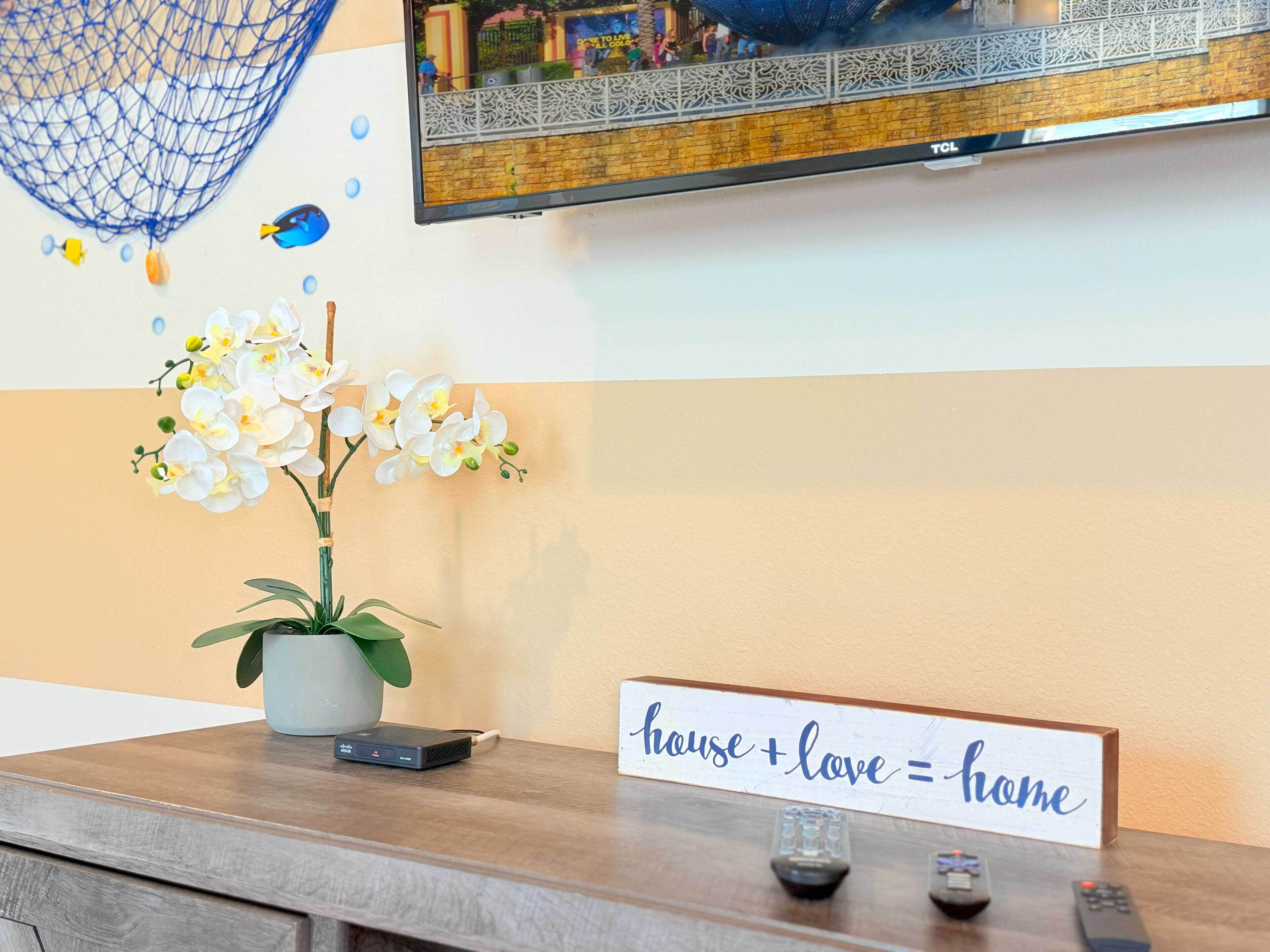 A wooden reception desk with a potted orchid, a sign reading "house + love = home", and a television mounted on the wall.