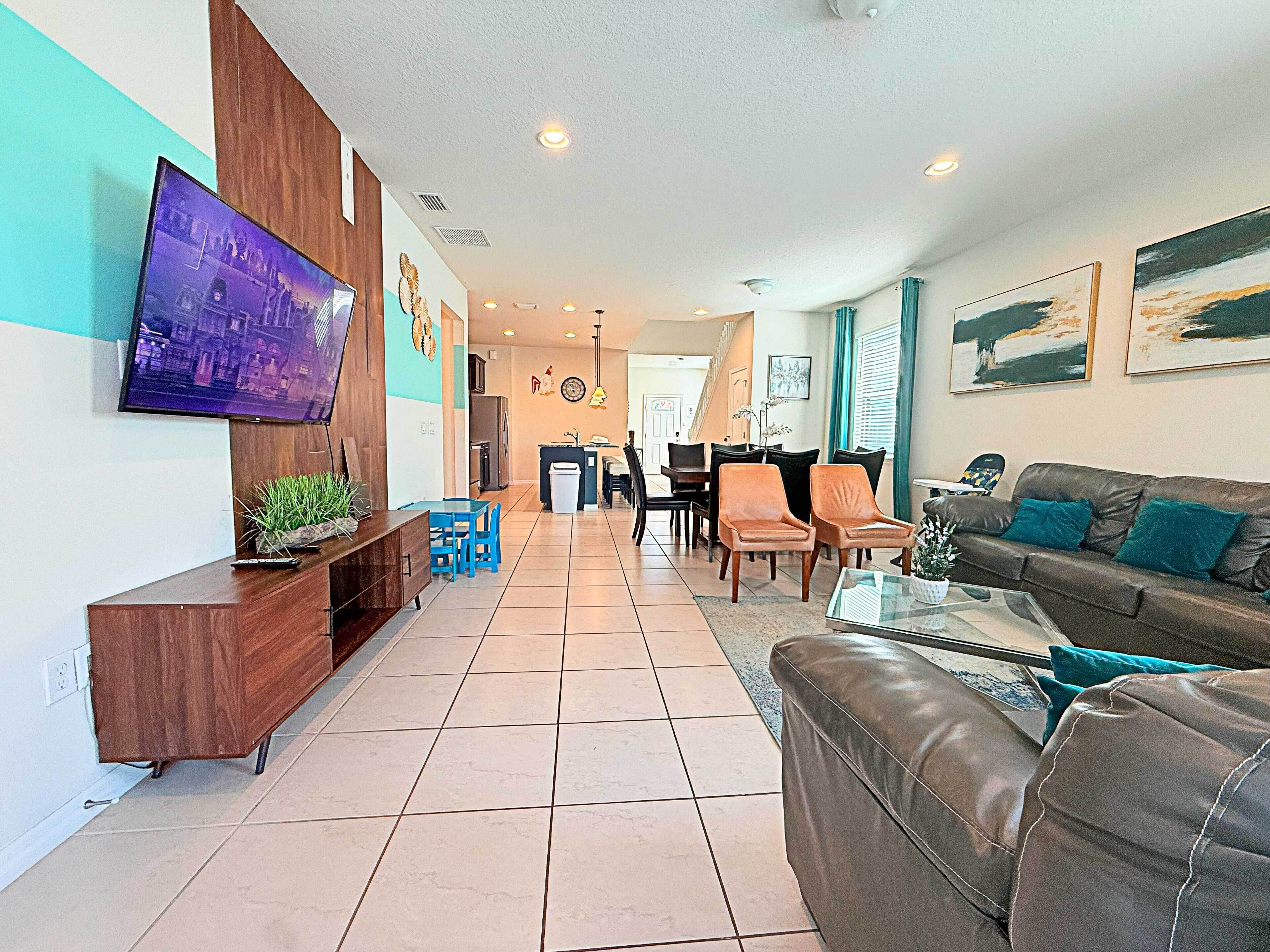 A modern living room with a flat-screen TV, a brown leather sofa, and a dining area with chairs and a table.