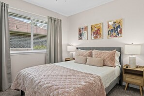 Ein Schlafzimmer mit einem Bett, zwei Nachttischen, einem Fenster mit Vorhängen und drei an der Wand hängenden Bildern.