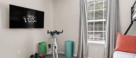 A room with a TV mounted on the wall, a bicycle, exercise equipment, and a window with curtains.