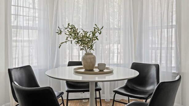 Une table de salle à manger ronde, ornée d’un vase de plantes vertes, entourée de chaises noires et recouverte d’une nappe blanche.