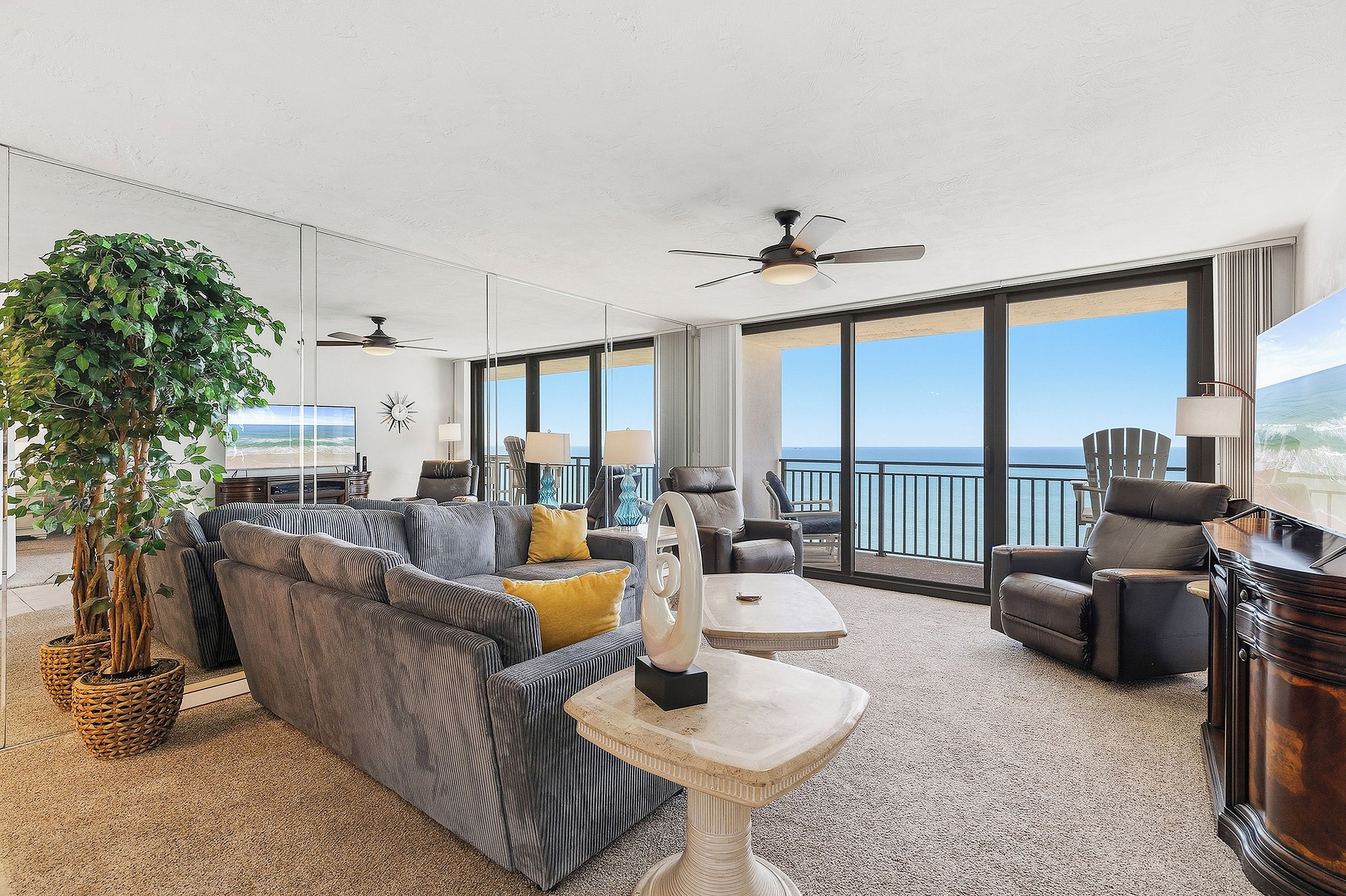 A spacious living area with a large sofa, armchairs, and a coffee table. There are ceiling fans, a large window with a view of the ocean, and a potted plant.