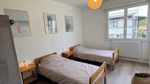 Une chambre avec deux lits simples, une table de chevet avec une lampe, un tapis tissé et une fenêtre donnant sur un immeuble et de la verdure.