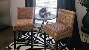 Een kamer met twee rieten stoelen, een kleine tafel, een tapijt met een zebraprint, een koelkast en een raam met gordijnen.