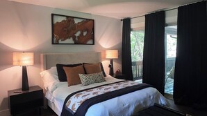Een slaapkamer met een groot bed, twee nachtkastjes met lampen, een rug met luipaardprint en uitzicht op het groen door een raam met gordijnen.