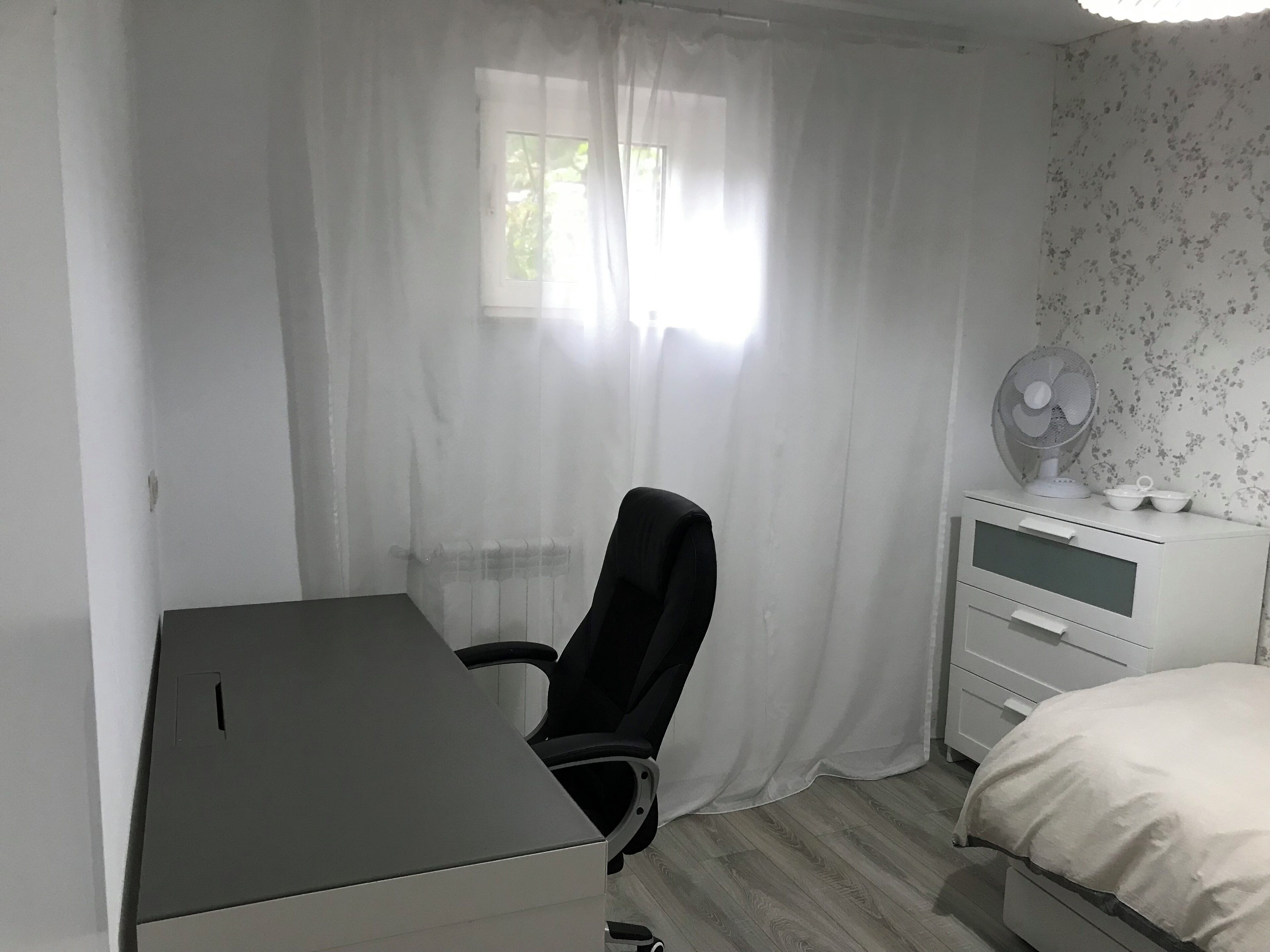 A bedroom with a bed, a desk, a chair, a nightstand, a fan, and a window with curtains.