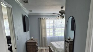 A bedroom with a wooden floor, a bed with a floral bedspread, a dresser, a mirror, and a ceiling fan. - A place to call home. (Foley)