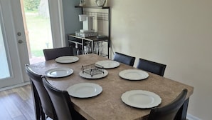 A dining room with a rectangular table set for six, surrounded by six chairs. There is a kitchen area with a countertop and a hanging rack above the sink. - A place to call home. (Foley)