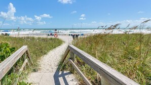 A wooden boardwalk leading to a beach with waves and umbrellas in the distance.