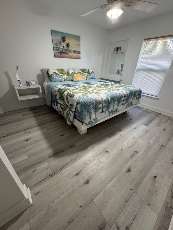 A bedroom with a bed, a nightstand, a ceiling fan, a window with blinds, and a painting on the wall. - Penguins on the Beach Villas (Cocoa Beach)