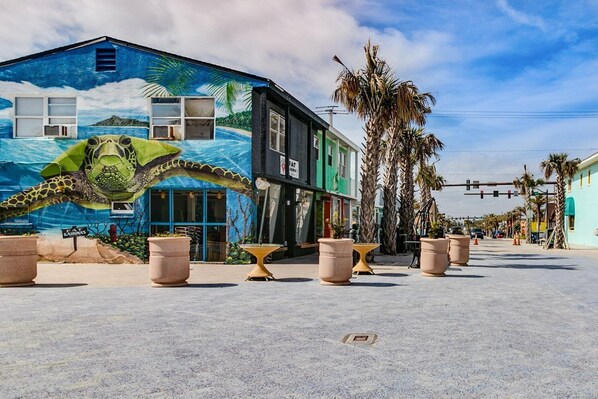 A street with a mural of a sea turtle on a building, palm trees, and outdoor seating areas. - Penguins on the Beach Villas (Cocoa Beach)