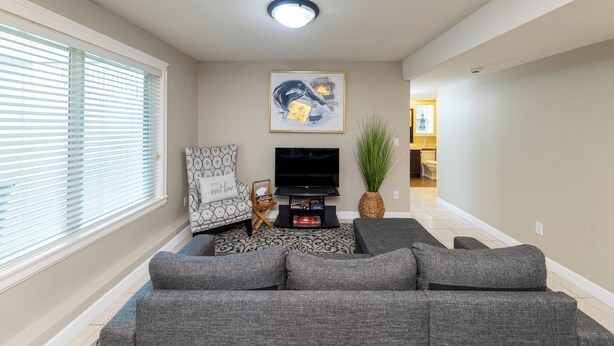A living room with a grey sectional sofa, a flat-screen TV, a painting, a potted plant, and a patterned rug. - Luxury 2 Bedroom Surrey Basement Suite, Sleeps 6 (Surrey)