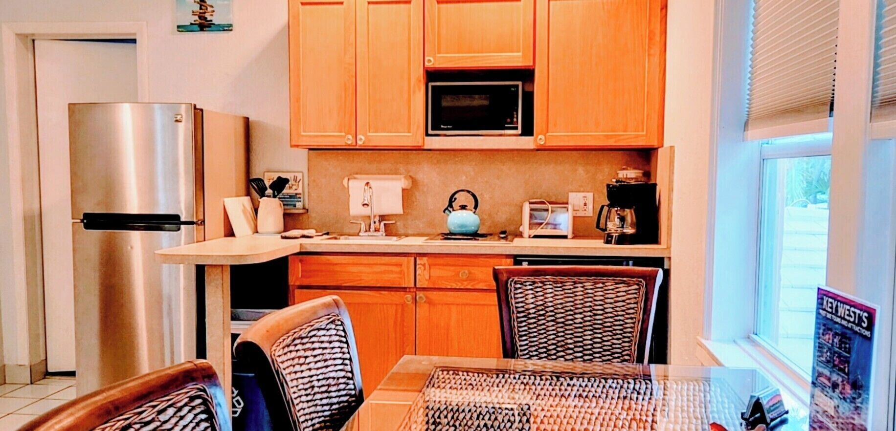 A compact kitchen with wooden cabinets, a stainless steel refrigerator, a microwave, and a dining area with wicker chairs.